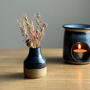 May include: A small, two-tone ceramic vase with dried flowers, in blue and tan. Beside it is a blue ceramic oil burner with a lit candle inside. The items are on a wooden surface, creating a warm, inviting atmosphere.