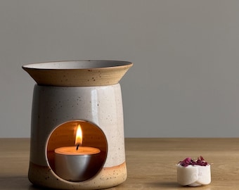 May include: A ceramic essential oil diffuser with a lit tea light candle inside. The diffuser is off-white with a speckled finish and a brown rim. A small white wax melt with dried rose petals sits nearby. The diffuser is on a wooden surface.