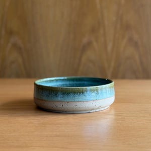May include: A shallow ceramic bowl with a low profile and straight sides. The interior features a deep teal glaze, while the exterior transitions from a vibrant turquoise band at the top to a speckled, off-white base at the bottom.
