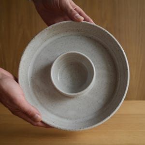 May include: A round ceramic serving dish with a speckled off-white glaze, featuring a wide outer rim and a smaller, integrated central bowl. The stoneware has a textured, earthy appearance with visible dark flecks throughout the surface.