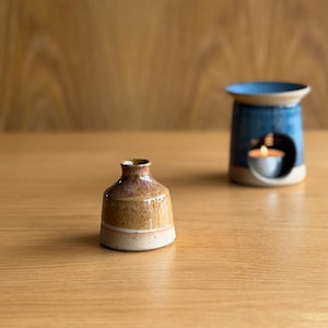 May include: A small, speckled ceramic bud vase with a tapered neck and a two-tone brown and cream glaze, positioned in front of a blue ceramic oil burner containing a lit tea light candle, both featuring a smooth, glossy finish.
