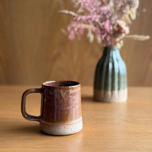May include: A ceramic mug with a glossy, mottled brown and amber glaze on the upper portion and a speckled, matte cream-coloured base. The mug features a sturdy, curved handle and a cylindrical body with a slightly tapered rim and a flat bottom.