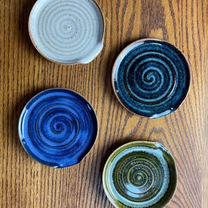 May include: Four ceramic spoon rests with a swirl design. One is white with brown speckles, one is blue with a dark blue swirl, one is dark blue with a light blue swirl, and one is green with a light green swirl.