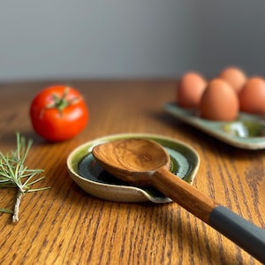 May include: A green and white ceramic spoon rest with a wooden spoon resting in it. The spoon has a dark grey handle.