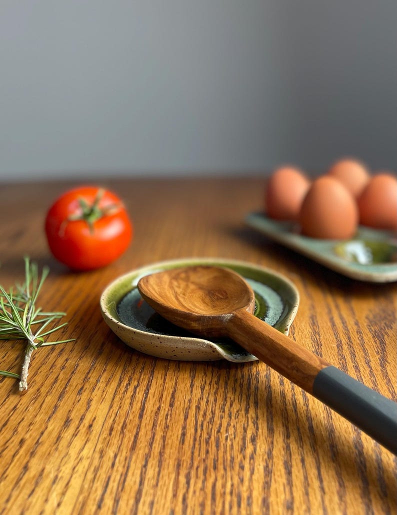 May include: A green and white ceramic spoon rest with a wooden spoon resting in it. The spoon has a dark grey handle.