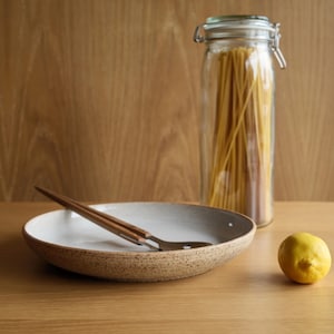 May include: A shallow, speckled beige ceramic bowl with a white interior, containing a metal spoon with a wooden handle. Next to the bowl is a clear glass jar filled with long spaghetti noodles and a single yellow lemon resting on the surface.