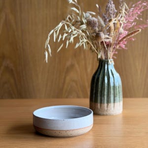 May include: A shallow, round ceramic bowl with a speckled white interior and a light tan base. Beside it stands a tall, fluted ceramic vase featuring a dark green glazed upper section and a textured, natural tan base filled with dried grasses.
