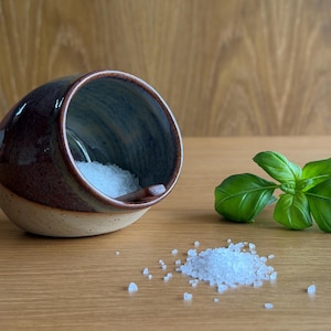 May include: A handcrafted ceramic salt pig with a deep brown glaze and a pale beige base. The interior is filled with coarse white salt. Fresh basil leaves are visible, suggesting culinary use.