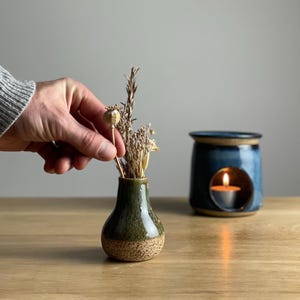 May include: A small, green and brown ceramic vase holds dried flowers. A blue ceramic oil warmer with a lit candle sits nearby. The vase is approximately 7.6 cm tall. The oil warmer has a circular opening.