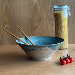 May include: A ceramic bowl with a blue rim and brown and tan textured exterior holds wooden chopsticks. A glass jar filled with spaghetti and a cluster of red cherry tomatoes are also on the wooden surface.