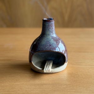 May include: A ceramic incense holder with a rounded body and a slender neck. The holder is glazed in blue, purple, and brown hues, with a cream base. Smoke rises from the top, indicating the incense is lit. A wooden incense stick is visible.