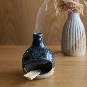 May include: A dark blue ceramic incense holder with a speckled light base and a wide opening. A piece of light-coloured wood is placed inside the opening, emitting a thin trail of white smoke that rises vertically from the top of the vessel.