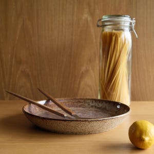 May include: A shallow, speckled brown ceramic bowl with a pair of wooden-handled utensils resting inside. Beside the bowl sits a clear glass jar filled with long spaghetti noodles and a single bright yellow lemon on the flat surface.