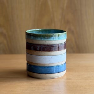 May include: A stack of four ceramic bowls with a speckled beige base. The top bowl is teal, the second is dark brown, the third is light blue, and the bottom bowl is a medium blue. Each bowl features a smooth, glazed interior and exterior rim.
