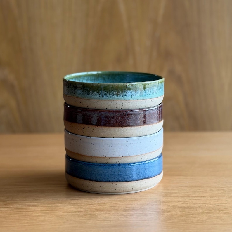 May include: A stack of four ceramic bowls with a speckled beige base. The top bowl is teal, the second is dark brown, the third is light blue, and the bottom bowl is a medium blue. Each bowl features a smooth, glazed interior and exterior rim.