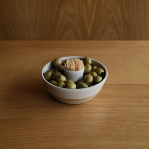 May include: A speckled ceramic bowl with a central cylindrical holder filled with wooden toothpicks, surrounded by a collection of pitted green olives. The bowl features a light cream-coloured glaze with dark flecks and a smooth, rounded base.