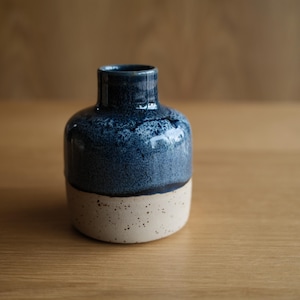 May include: A small ceramic vase with a rounded body and a narrow neck. The upper portion features a deep, glossy blue glaze with a mottled texture, while the lower portion is a matte, speckled off-white base with a distinct horizontal transition.