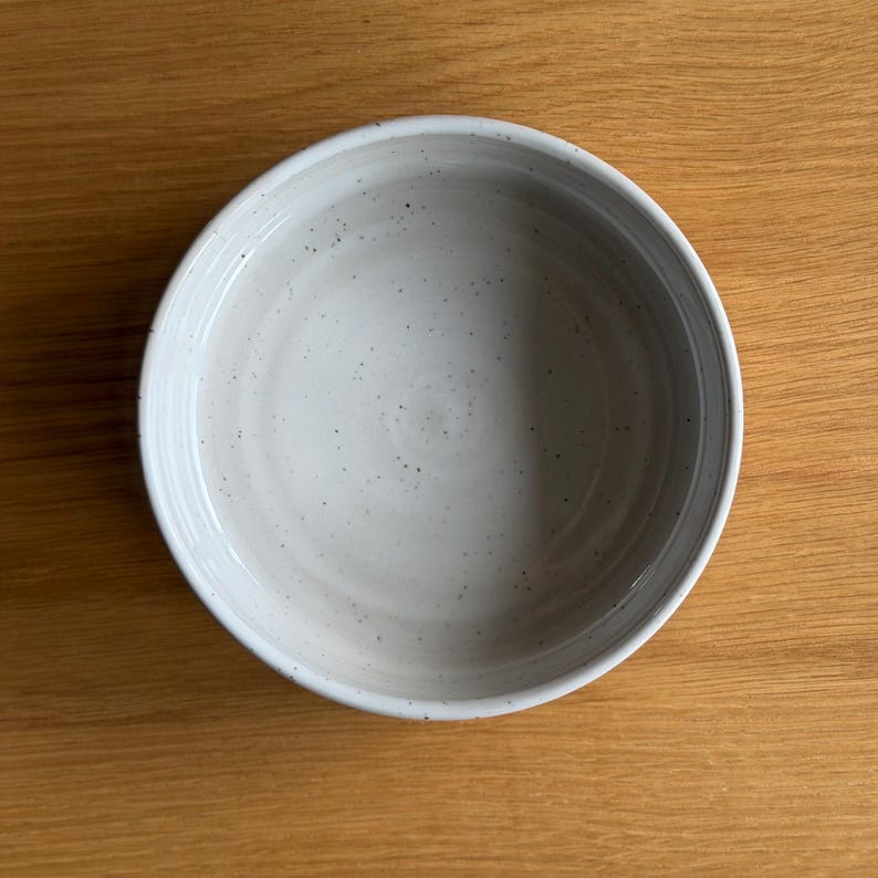 May include: A round ceramic bowl with a light gray, speckled finish. The bowl features a smooth, slightly concave interior surface with visible dark gray flecks distributed across the light gray glaze, creating a subtle, textured appearance.
