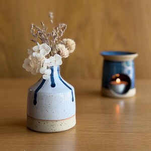 May include: A small ceramic bud vase with a speckled light blue and white glaze featuring dark blue drip patterns, containing a bouquet of dried white hydrangea and beige flowers, with a matching blue ceramic oil burner in the background.