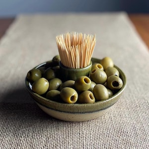 May include: A green ceramic bowl filled with green olives and a small green ceramic cup filled with wooden toothpicks.