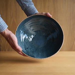 May include: A large, round ceramic bowl featuring a deep blue, speckled glaze with a smooth, glossy finish. The interior of the bowl displays a gradient of dark blue tones, while the outer rim has a light, speckled, natural-coloured edge.