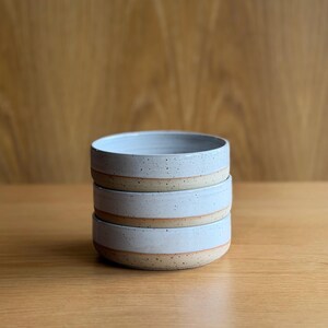 May include: A stack of three round ceramic bowls featuring a light blue glazed interior and upper exterior, transitioning to a speckled beige unglazed base. Each bowl has a low profile with straight sides and a smooth, consistent circular rim.