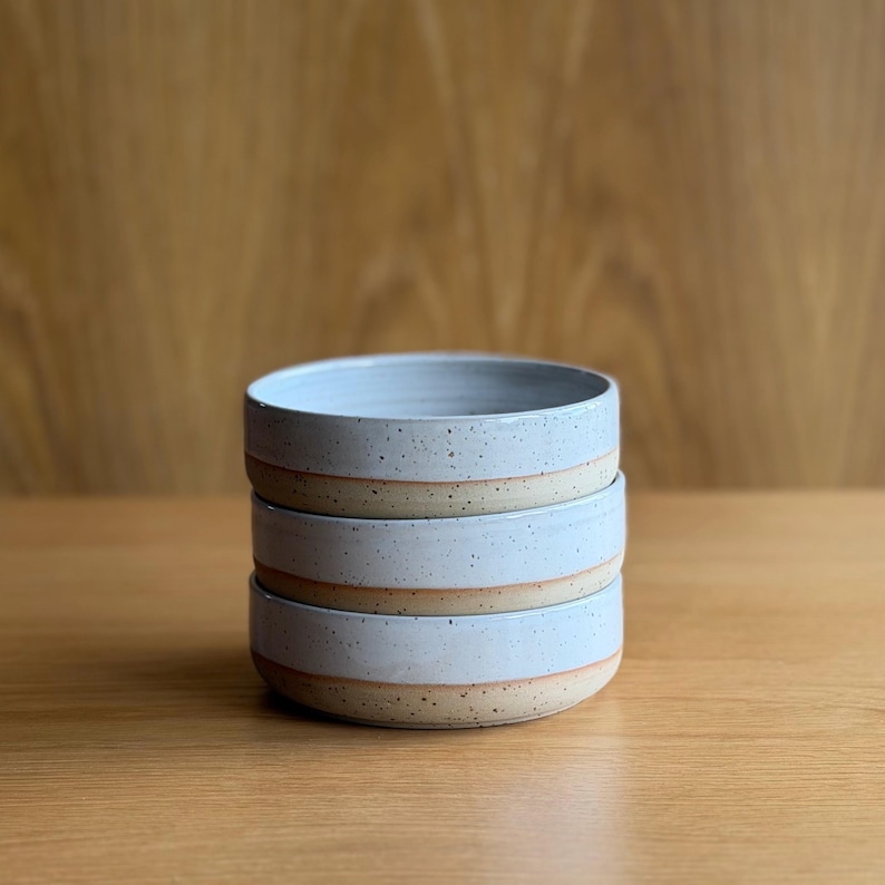May include: A stack of three round ceramic bowls featuring a light blue glazed interior and upper exterior, transitioning to a speckled beige unglazed base. Each bowl has a low profile with straight sides and a smooth, consistent circular rim.