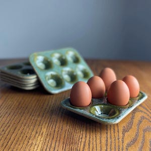 May include: A stack of ceramic egg holders in a light green hue, with a matching tray holding five brown eggs. The holders have six circular indentations. The items are on a wooden surface.