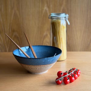 May include: A ceramic bowl with a deep blue glazed interior and rim, transitioning to a speckled beige base. Two wooden chopsticks rest inside the bowl. Beside it sits a clear glass jar filled with spaghetti and a vine of small red cherry tomatoes.
