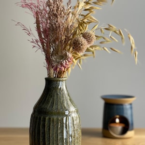 May include: A green ceramic vase with vertical grooves, filled with dried flowers in pink, brown, and beige. A blue and tan candle warmer is to the right. The vase has a speckled tan base.