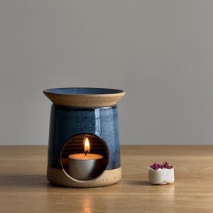 May include: A blue ceramic essential oil diffuser with a tan rim and base. A lit tealight candle is inside the diffuser, and a small white wax melt with dried flowers sits beside it. The diffuser is designed to hold essential oils.