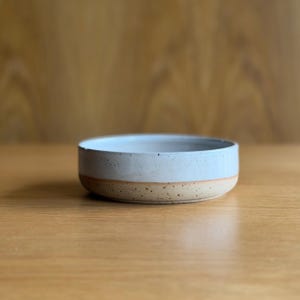 May include: A round ceramic bowl featuring a two-tone design with a smooth, light blue glazed upper section and a speckled, unglazed tan base. The bowl has a low profile with straight sides and a flat rim, showcasing a handcrafted aesthetic.