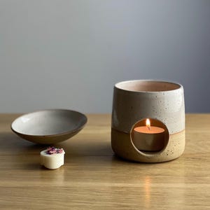 May include: A white ceramic wax melt warmer with a speckled glaze. The warmer has a round opening in the centre to hold a tea light candle. A small white wax melt with dried flowers is in the foreground.