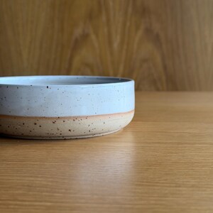 May include: A shallow, round ceramic bowl featuring a two-tone design. The upper portion is finished in a smooth, glossy light blue glaze with dark speckles, while the lower base remains in an unglazed, textured, speckled beige clay finish.