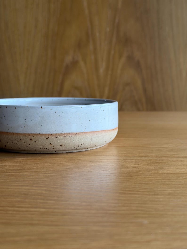 May include: A shallow, round ceramic bowl featuring a two-tone design. The upper portion is finished in a smooth, glossy light blue glaze with dark speckles, while the lower base remains in an unglazed, textured, speckled beige clay finish.