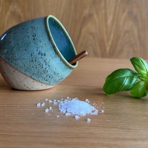 May include: A handmade ceramic salt cellar with a turquoise glaze and a wooden spoon. The salt cellar is tilted, with a small pile of coarse salt and fresh basil leaves on a wooden surface. The salt cellar is approximately 10 cm wide.