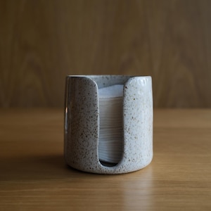 May include: A cylindrical ceramic holder with a speckled off-white finish and a vertical cutout opening. The container is filled with a stack of round white cotton pads, which are partially visible through the front opening of the ceramic vessel.