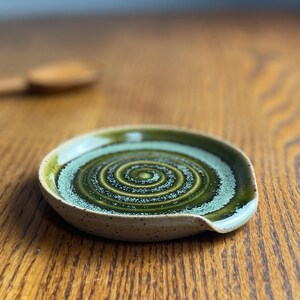 May include: A green and white ceramic spoon rest with a swirl design. The rest has a speckled glaze and a slight lip on one side.