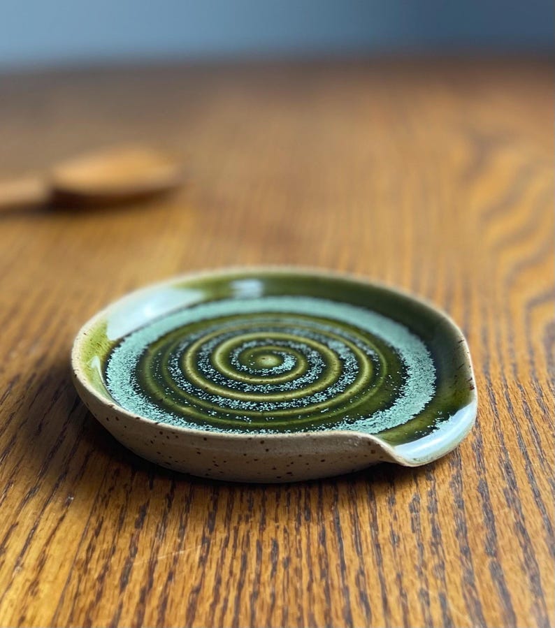 May include: A green and white ceramic spoon rest with a swirl design. The rest has a speckled glaze and a slight lip on one side.