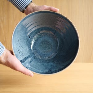 May include: A large, round ceramic bowl featuring a deep blue and charcoal grey speckled glaze. The interior surface displays a gradient of dark blue tones with a textured, mottled finish, while the outer rim is a light, neutral beige colour.