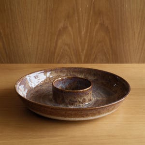 May include: A round ceramic serving dish with a central integrated bowl, featuring a mottled glaze in shades of brown, tan, and hints of blue. The surface has a glossy finish with visible variations in colour and texture across the entire piece.