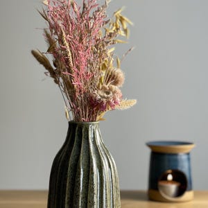 May include: A green ceramic vase with vertical ridges contains a bouquet of dried flowers in pink and beige tones. A blue and beige oil diffuser with a lit candle is in the background. The objects are on a wooden surface.