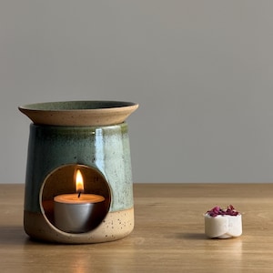 May include: A teal and tan ceramic oil diffuser with a lit tea light candle inside. A small white wax melt with dried rose petals is next to it. The diffuser has a round opening and a dish on top. The measurements are in centimeters.