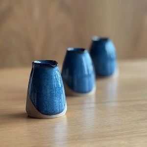 May include: Three ceramic vases with a blue glaze and a natural, unglazed base. The vases have an asymmetrical shape and are arranged on a wooden surface. The blue glaze has a speckled appearance, adding visual interest to the handmade pottery.