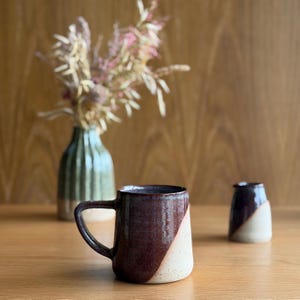 May include: A ceramic mug with a dark brown, glossy glaze on the upper portion and a speckled, off-white matte finish on the lower section, featuring a sturdy handle. A small matching creamer and a fluted green vase are positioned nearby.