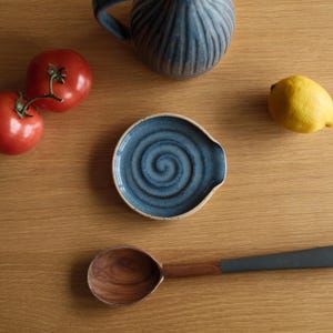 May include: A round ceramic spoon rest featuring a blue glaze with a dark spiral pattern in the centre and a light-coloured rim. The spoon rest is positioned near two red tomatoes, a yellow lemon, a blue ribbed pitcher, and a wooden spoon with a grey handle.