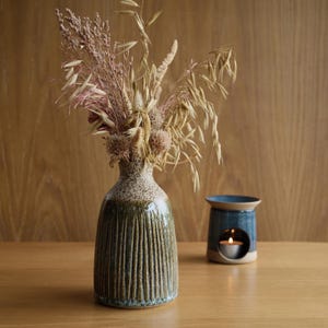 May include: A ceramic vase with a speckled cream neck and a vertically ribbed, dark green glazed body, filled with a bouquet of dried grasses and pink flowers. Beside it sits a small, blue ceramic oil burner with a lit tea light candle inside.