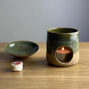 May include: A green and beige ceramic wax melt burner with a small white tea light candle burning inside. The burner has a hole in the centre for the candle and a small dish on the side for wax melts.