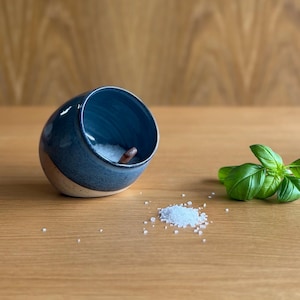 May include: A blue ceramic salt pig with a wooden spoon inside, on a wooden surface. A small pile of salt crystals and fresh basil leaves are next to the salt pig. The salt pig has a two-tone design with a light beige bottom.
