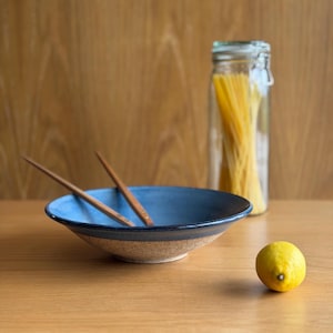 May include: A ceramic bowl with a deep blue interior and a speckled beige exterior, containing two wooden chopsticks. Beside the bowl sits a clear glass jar filled with long pasta strands and a single bright yellow lemon on the surface.